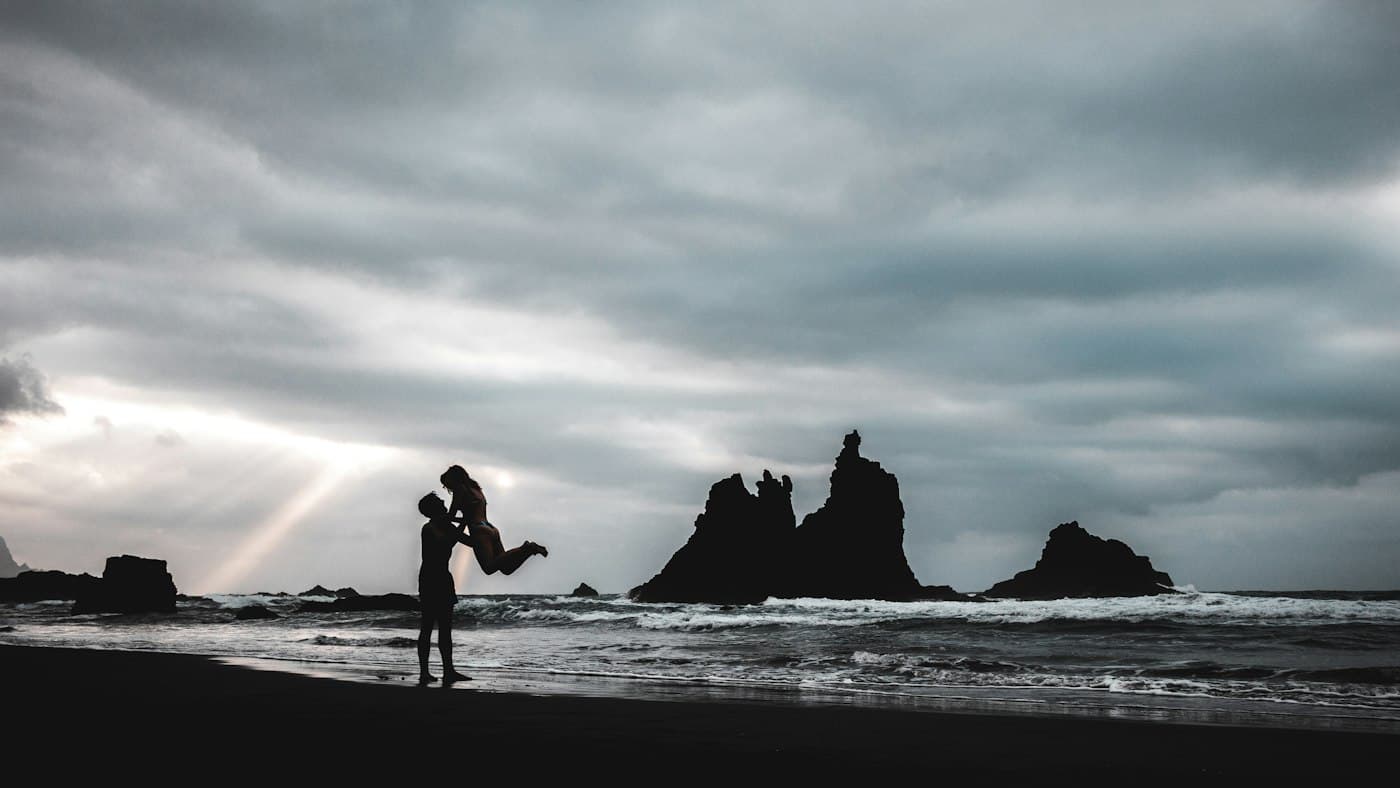 Um casal em silhueta numa praia de areia negra em Tenerife, com rochedos no mar atrás