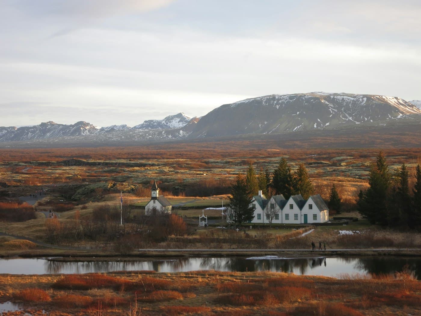 Die Kirche von Þingvellir und die weißen Giebel des alten Hofs zwischen rostrotem Gestrüpp, dahinter schneegestreifte Berge