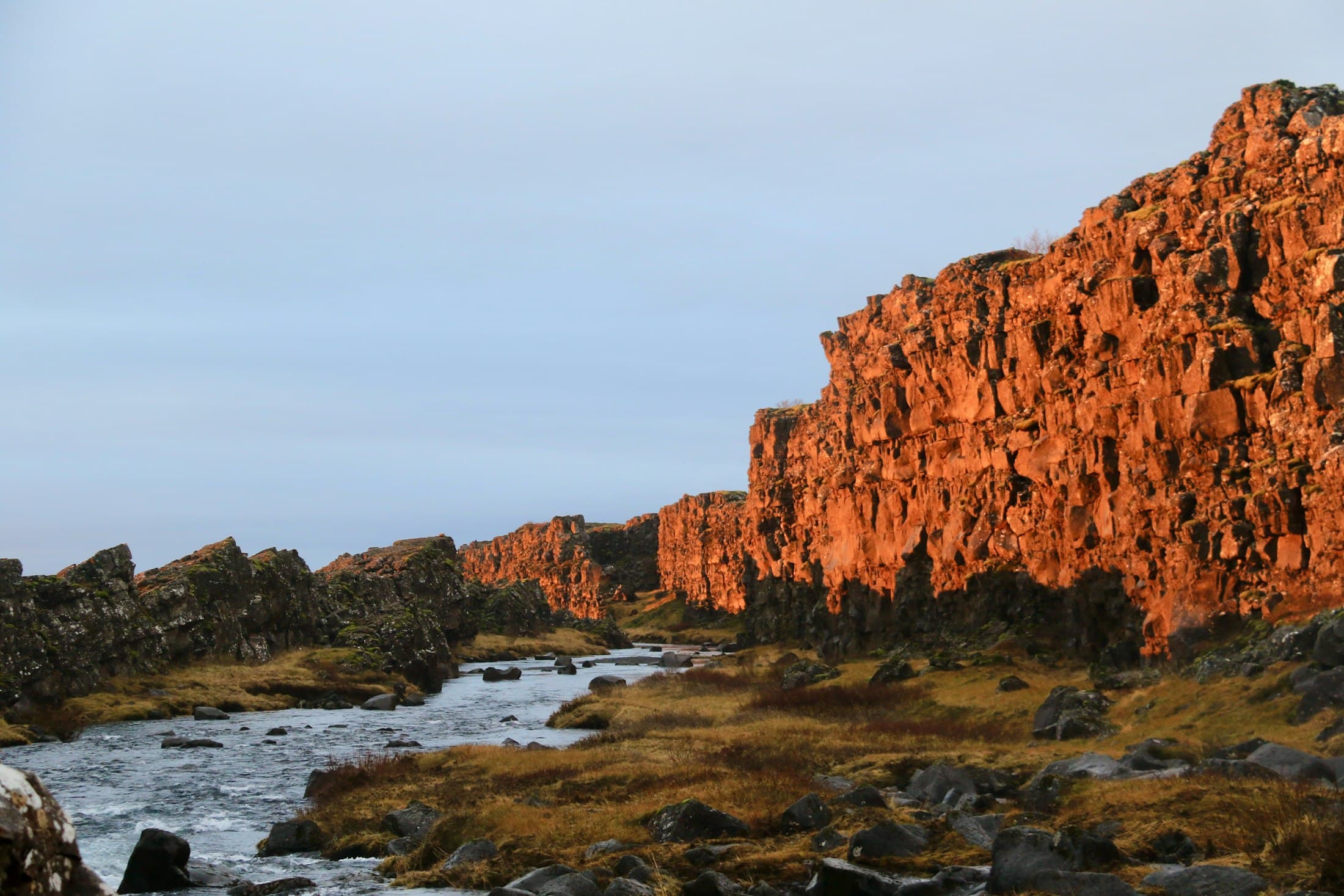 Las paredes de basalto de la falla de Þingvellir, encendidas de naranja por el sol bajo, con un río que cruza el cañón