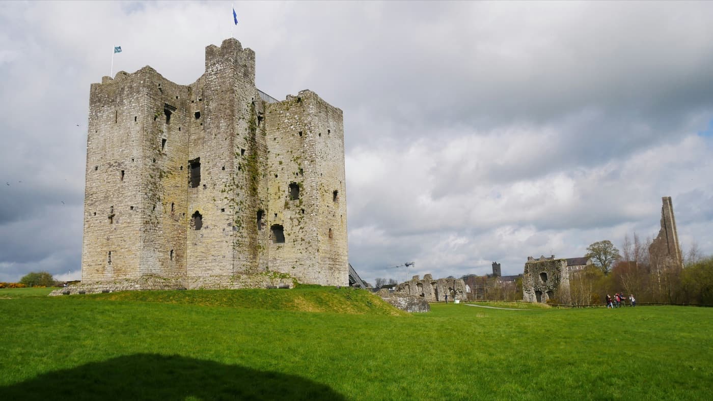The great stone keep of Trim Castle in County Meath on its grassy mound under a cloudy sky, with ruined curtain walls and the tall Yellow Steeple beyond the lawn