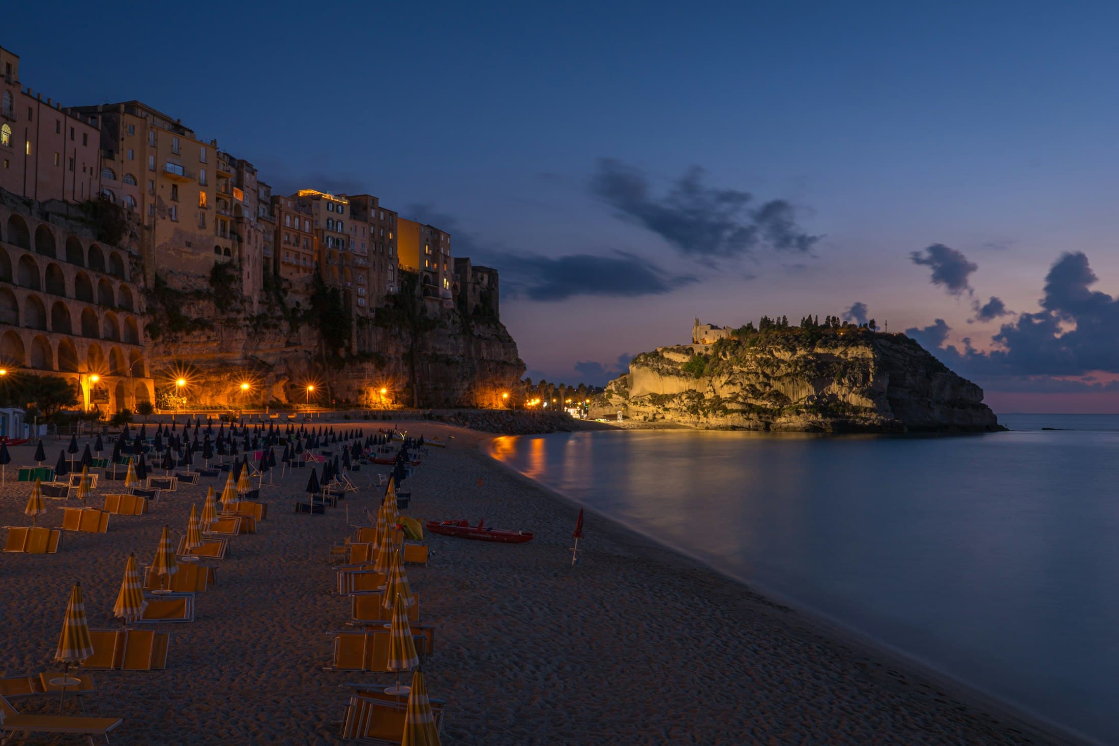 Tropea in der Abenddämmerung, die beleuchteten Häuser auf der Klippe über einem Strand und die Kirche Santa Maria dell’Isola auf ihrem Felsen