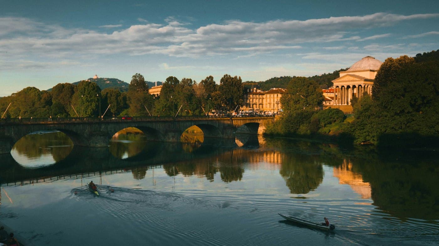 Der Po in Turin bei Sonnenuntergang: eine Steinbrücke, zwei Ruderer auf dem Wasser und die Kuppel der Gran Madre zwischen den Bäumen