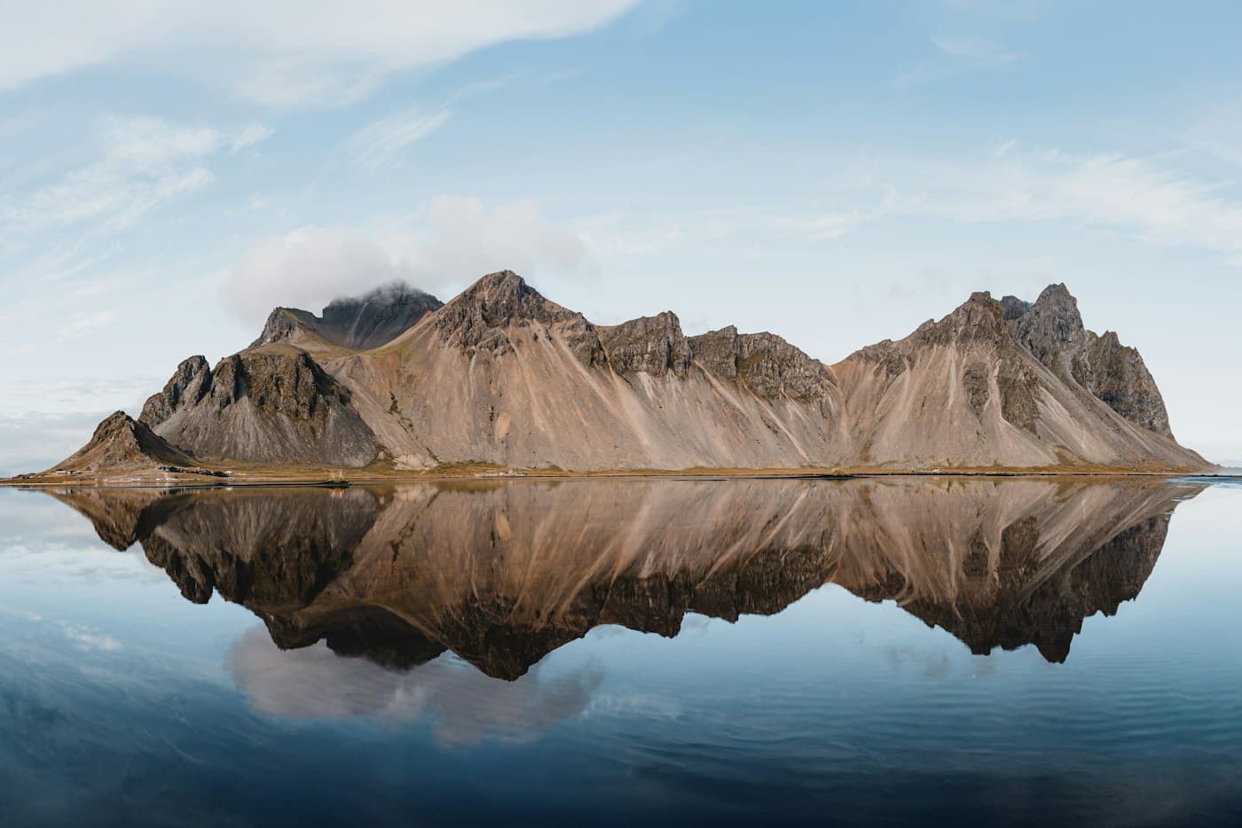 Der zackige Grat des Vestrahorn bei Stokksnes spiegelt sich in ruhigem, flachem Wasser unter hellem Himmel