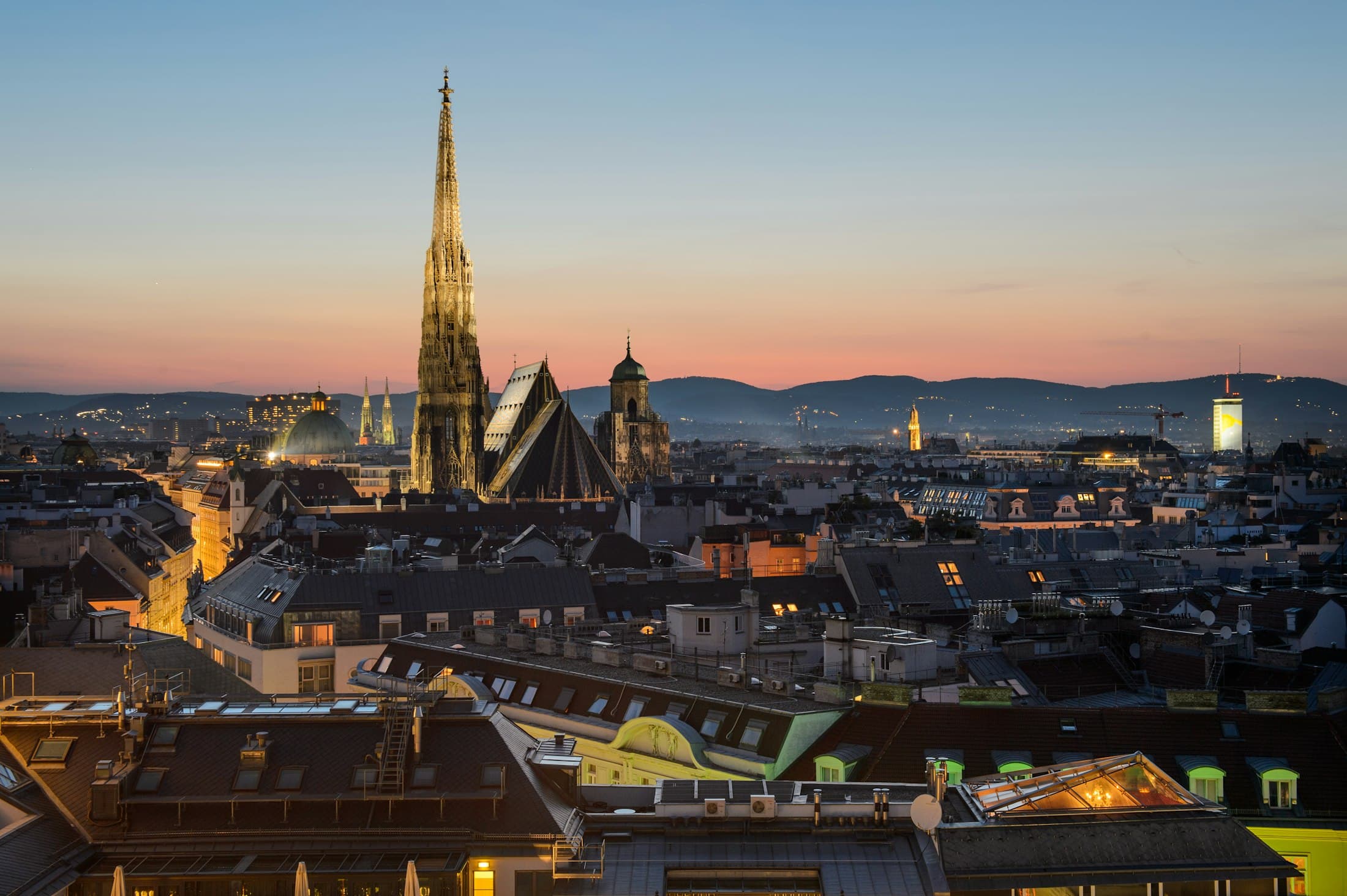 Der Turm des Stephansdoms über den Dächern Wiens in der Abenddämmerung, dahinter die Hügel des Wienerwalds