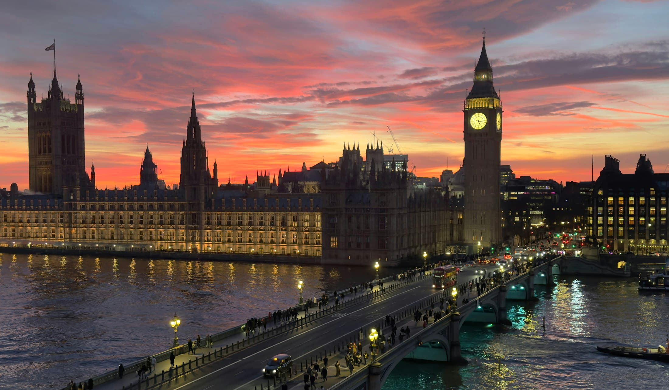 Das Parlament und das erleuchtete Zifferblatt des Elizabeth Tower vor einem rot-violetten Abendhimmel, davor die Westminster Bridge mit ihren Laternen und abendlichen Spaziergängern und die Lichter der Stadt, die sich in der Themse spiegeln
