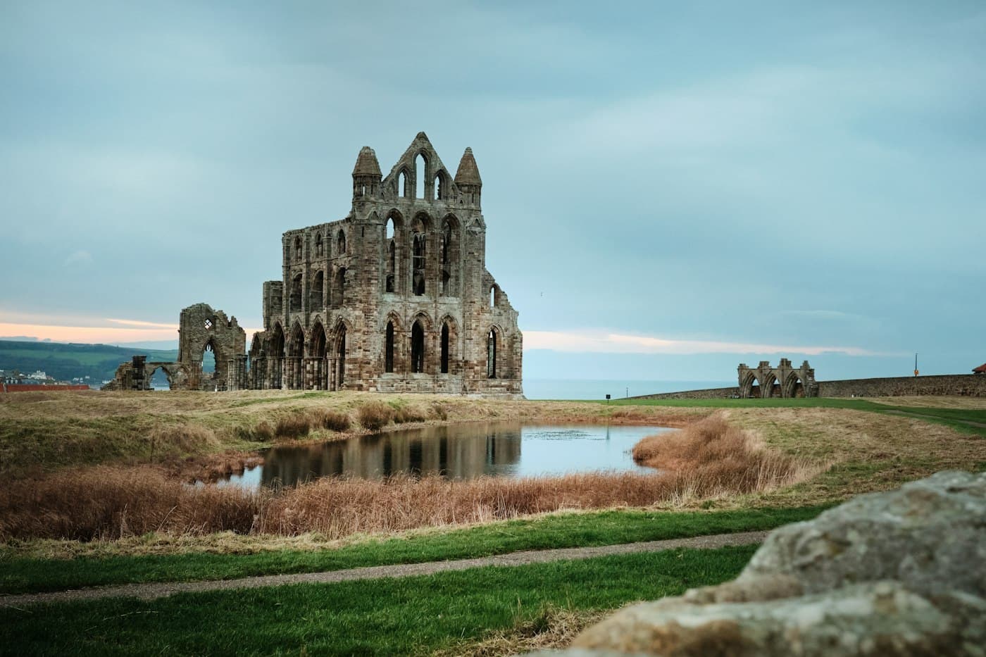 Die gotische Kirchenruine von Whitby Abbey, gespiegelt in einem von trockenem Schilf gesäumten Teich, am Horizont das Meer