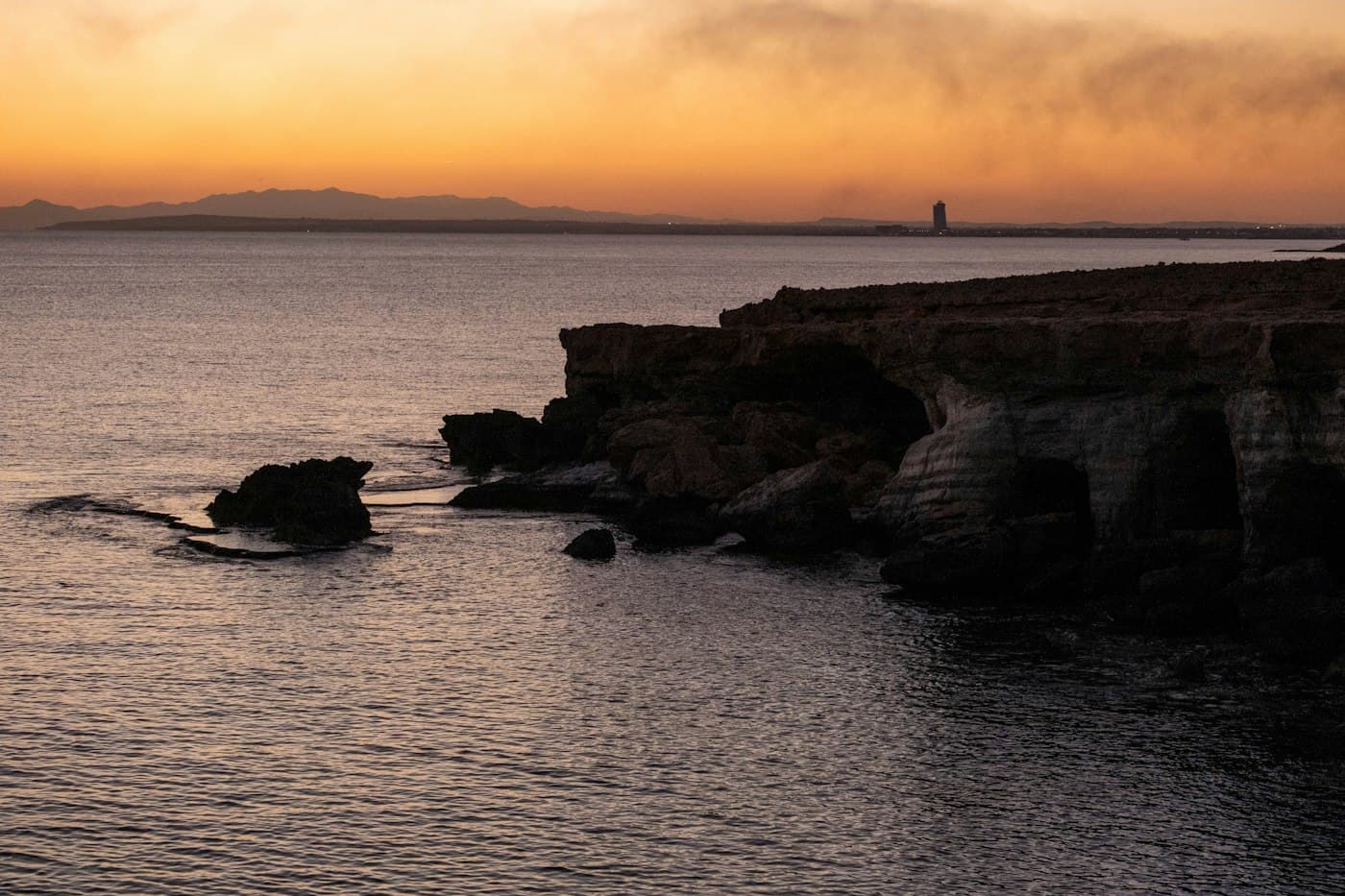 Sunset over the sea at Ayia Napa, the cliffs and sea caves in silhouette and hills faint on the horizon