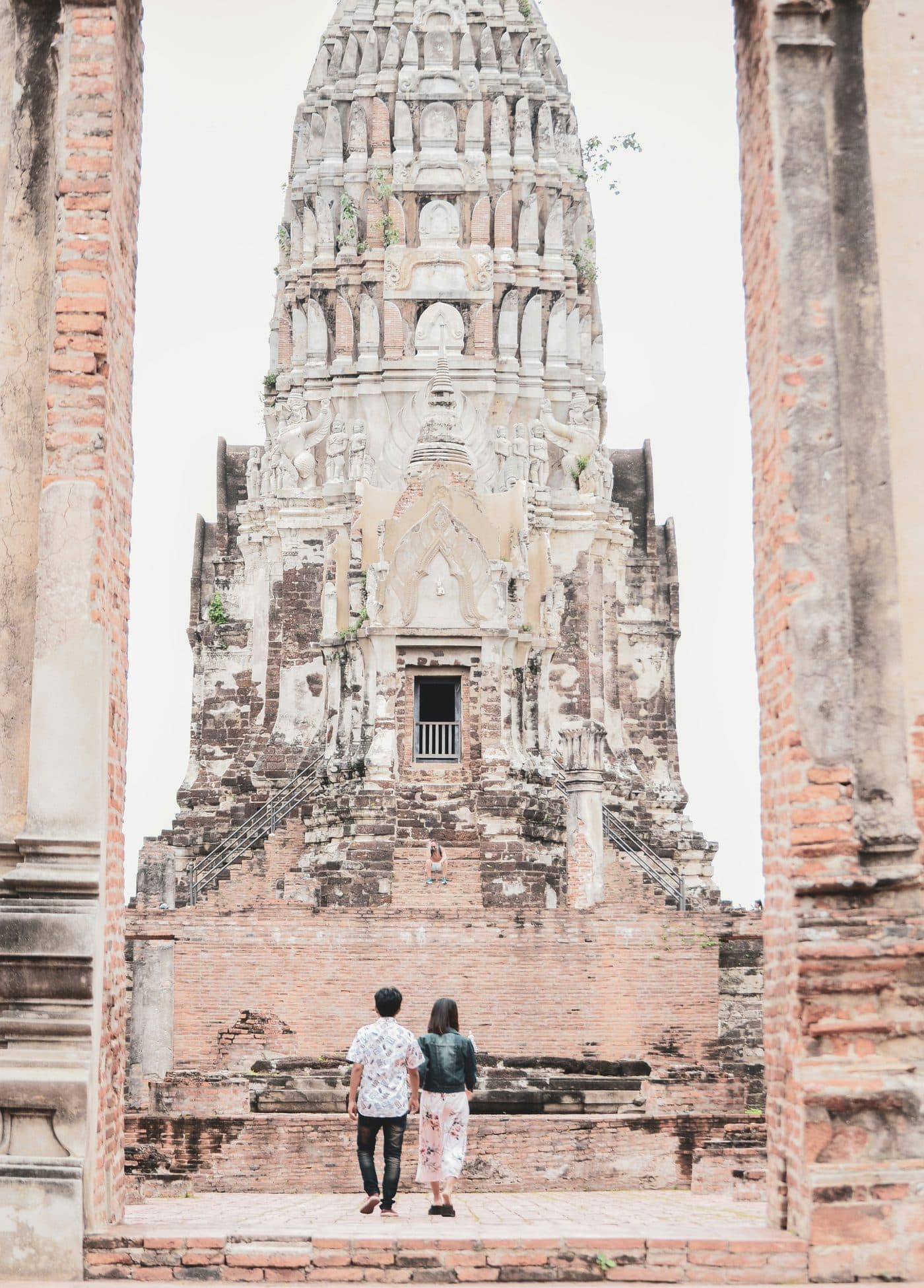 Ein Paar, von hinten gesehen, geht auf den großen verzierten Prang einer Tempelruine in Ayutthaya zu, eingerahmt von zwei verwitterten Backsteinpfeilern