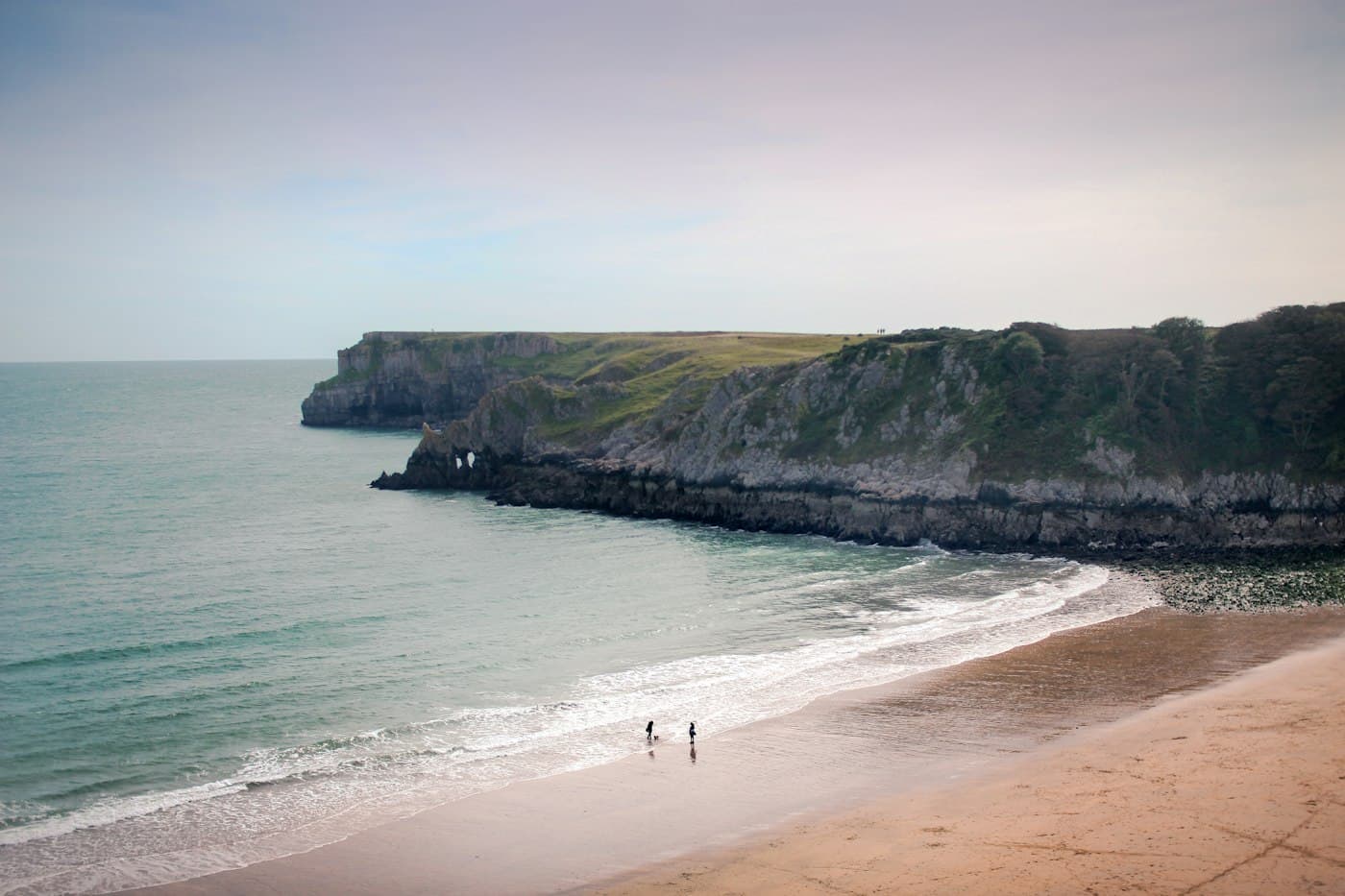 Barafundle Bay from above: a curve of pale sand and surf below a headland of grey cliffs with a small rock arch, and two people and a dog at the water’s edge