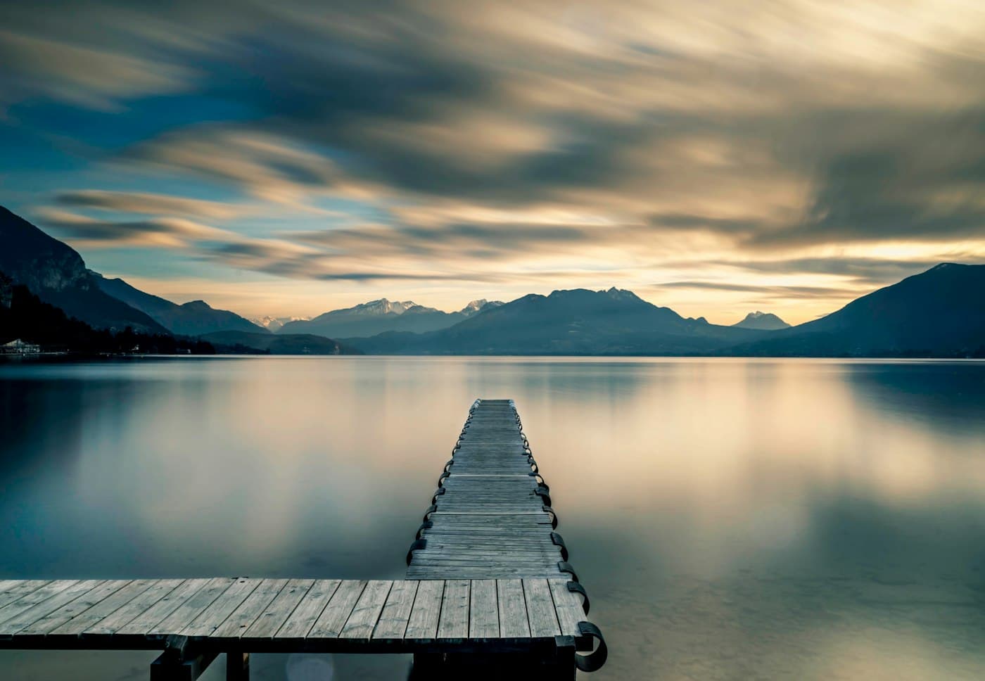A wooden jetty running out into the still water of Lake Annecy, mountains on the horizon under a long-exposure sky