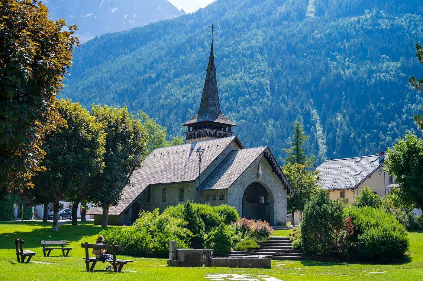 The stone church at Les Praz de Chamonix with its tall dark spire, a lawn with benches in front and forested slopes behind