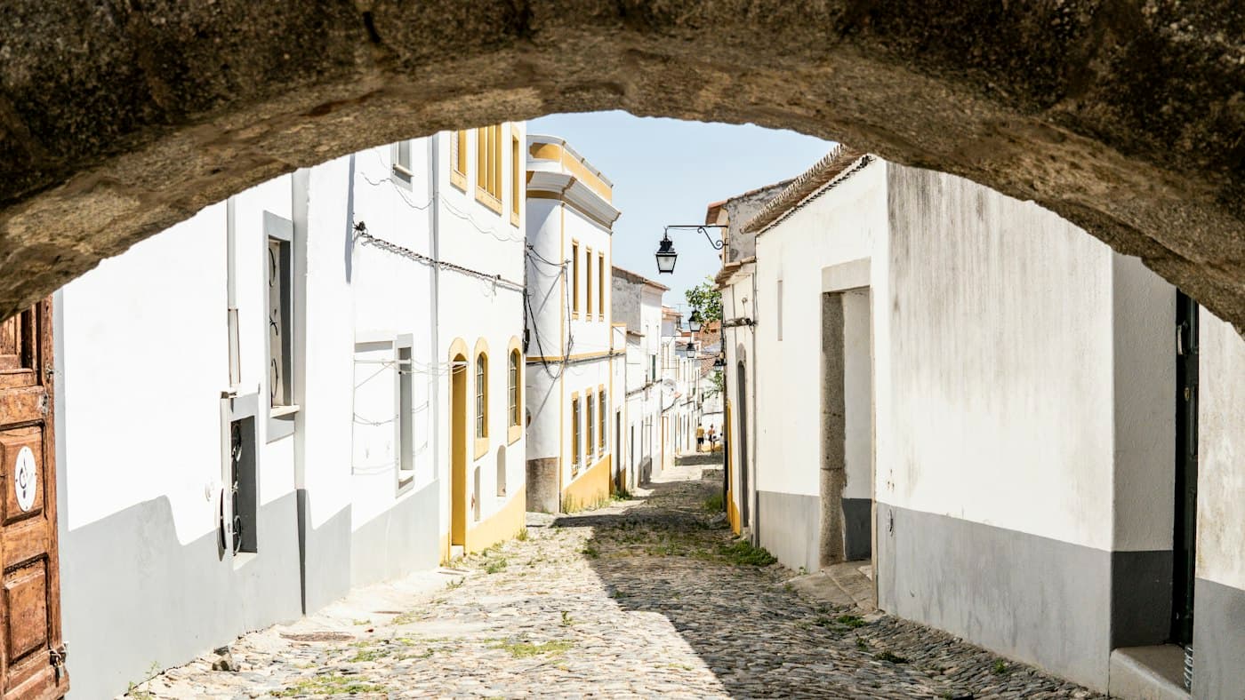 Uma rua empedrada de casas brancas com barras amarelas, vista através de um arco de pedra em Évora