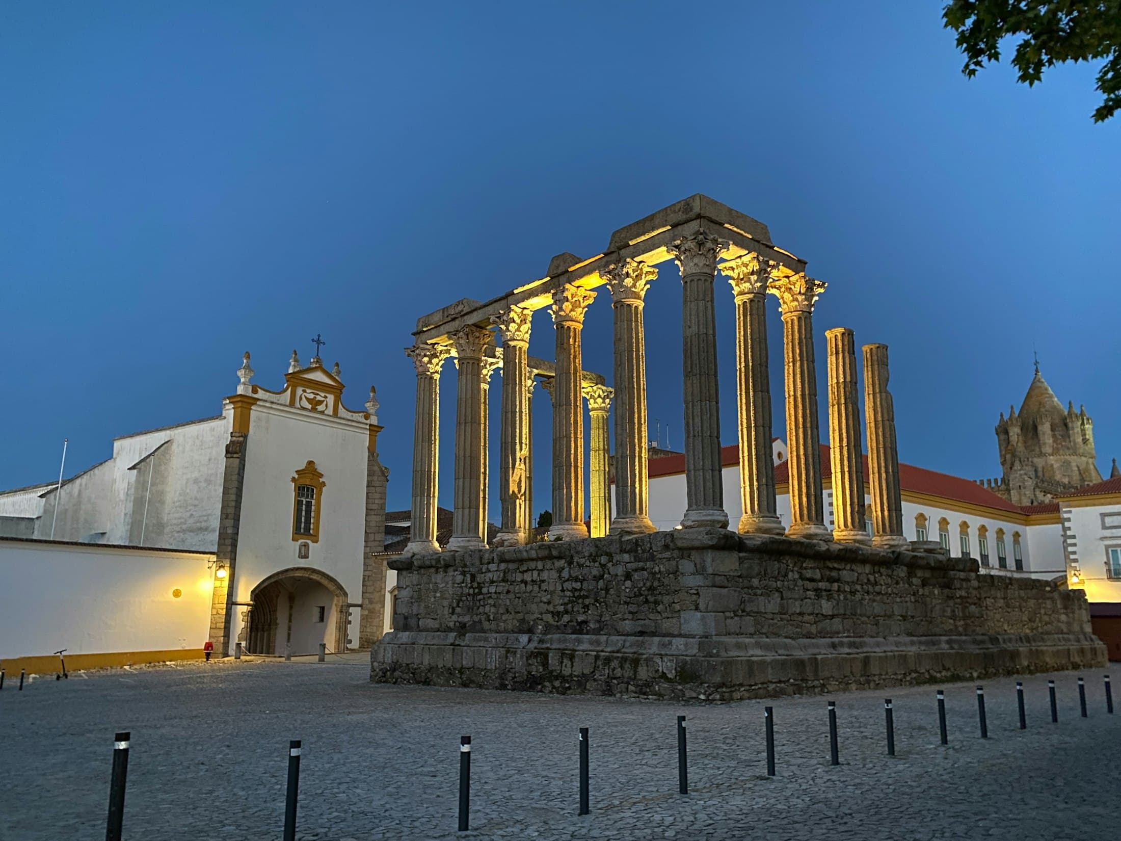 O templo romano de Évora iluminado ao anoitecer, com a igreja branca dos Lóios ao lado e a torre da Sé por trás