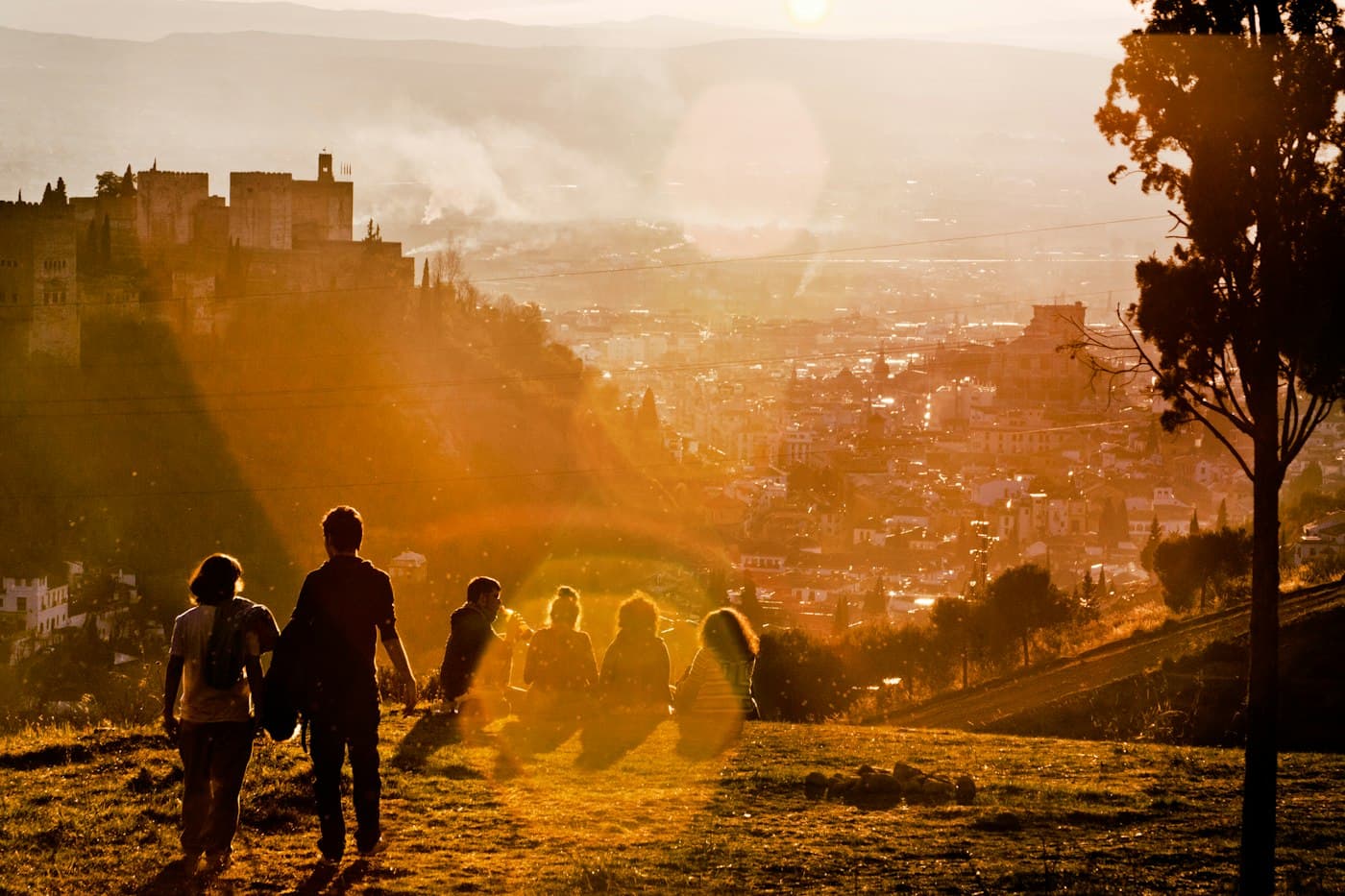 Sunset on the Sacromonte hill: a couple walks hand in hand past people sitting in the grass, the Alhambra’s towers on the left above the city