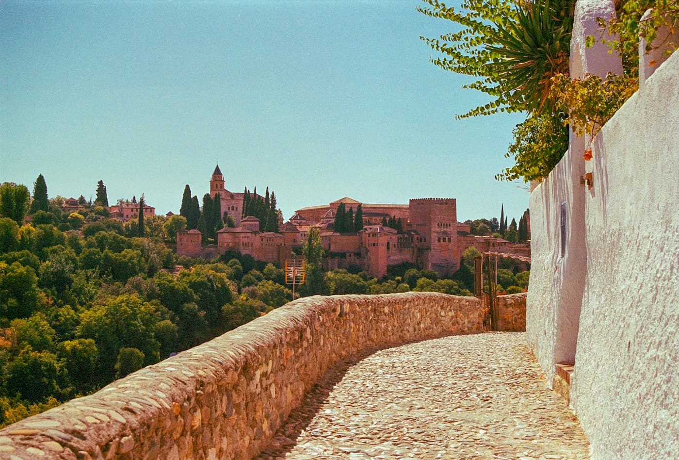 A cobbled lane beside a whitewashed wall in Granada, looking across the valley to the Alhambra