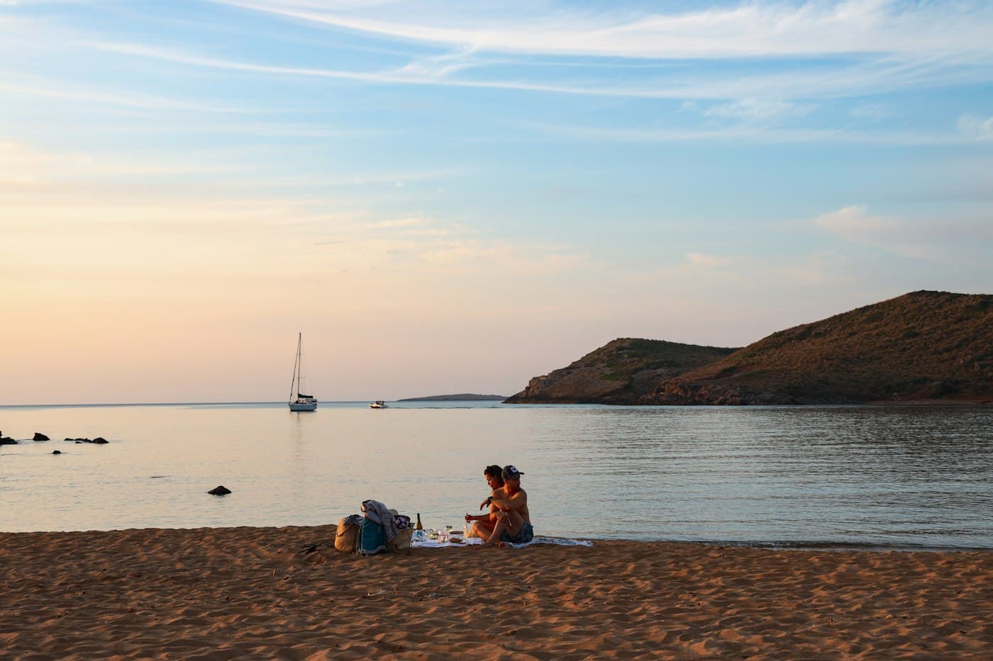 Una pareja sentada en la arena de una playa tranquila de Menorca al caer la tarde, con un velero fondeado en la bahía