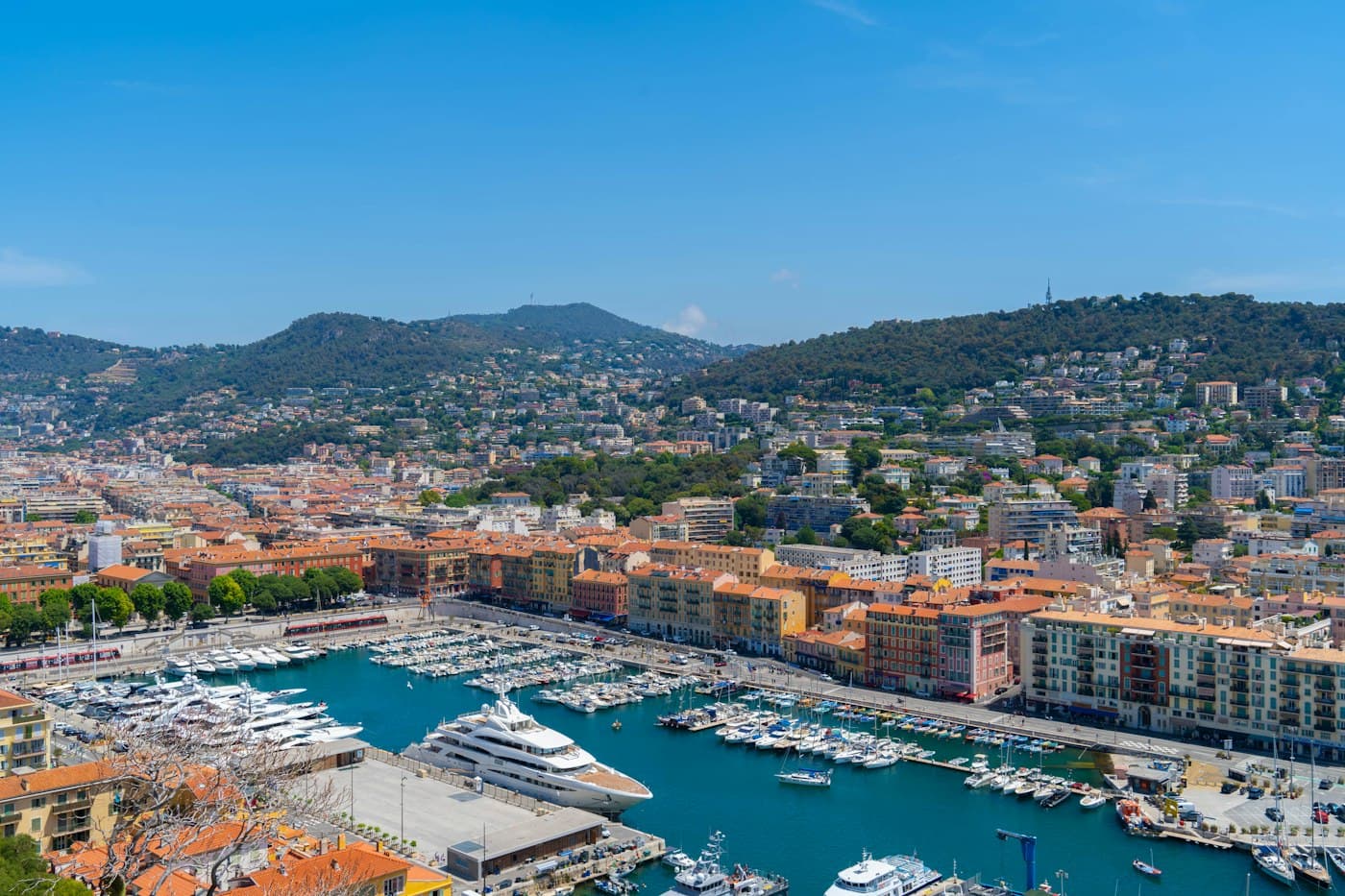 Port Lympia seen from above, boats along the quays, ochre and red buildings and green hills behind under a clear sky
