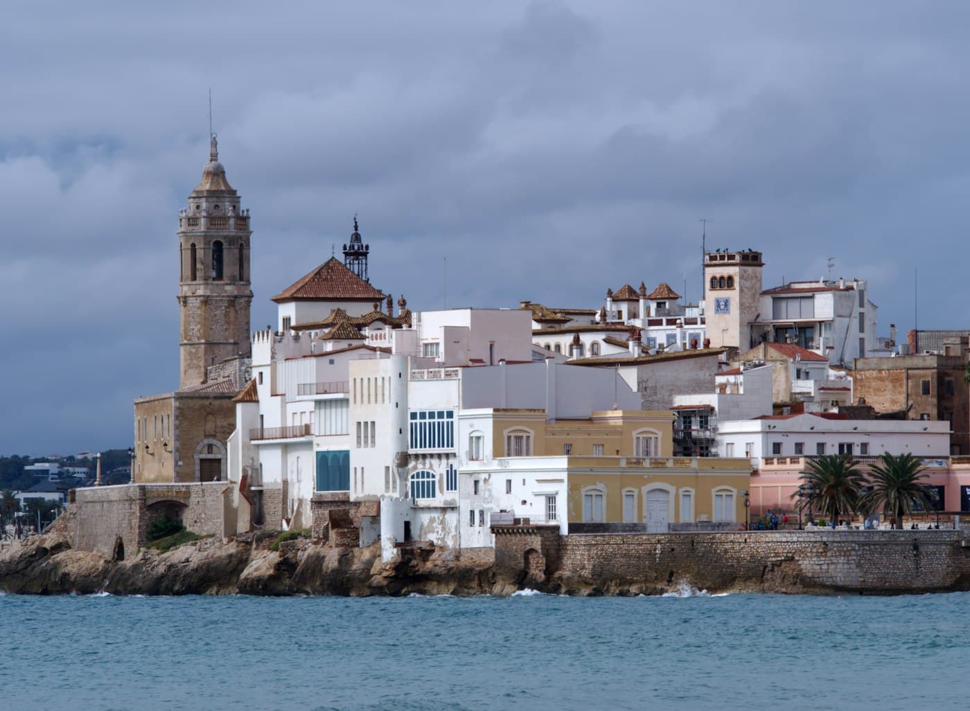 La iglesia de Sant Bartomeu i Santa Tecla y los edificios blancos de su lado sobre la punta rocosa de Sitges, vistos desde el mar bajo un cielo nublado