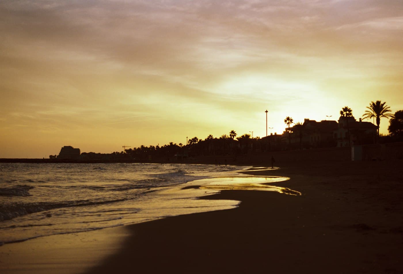 Atardecer sobre una playa de Sitges, la arena mojada brillando dorada y palmeras a contraluz a lo largo del paseo