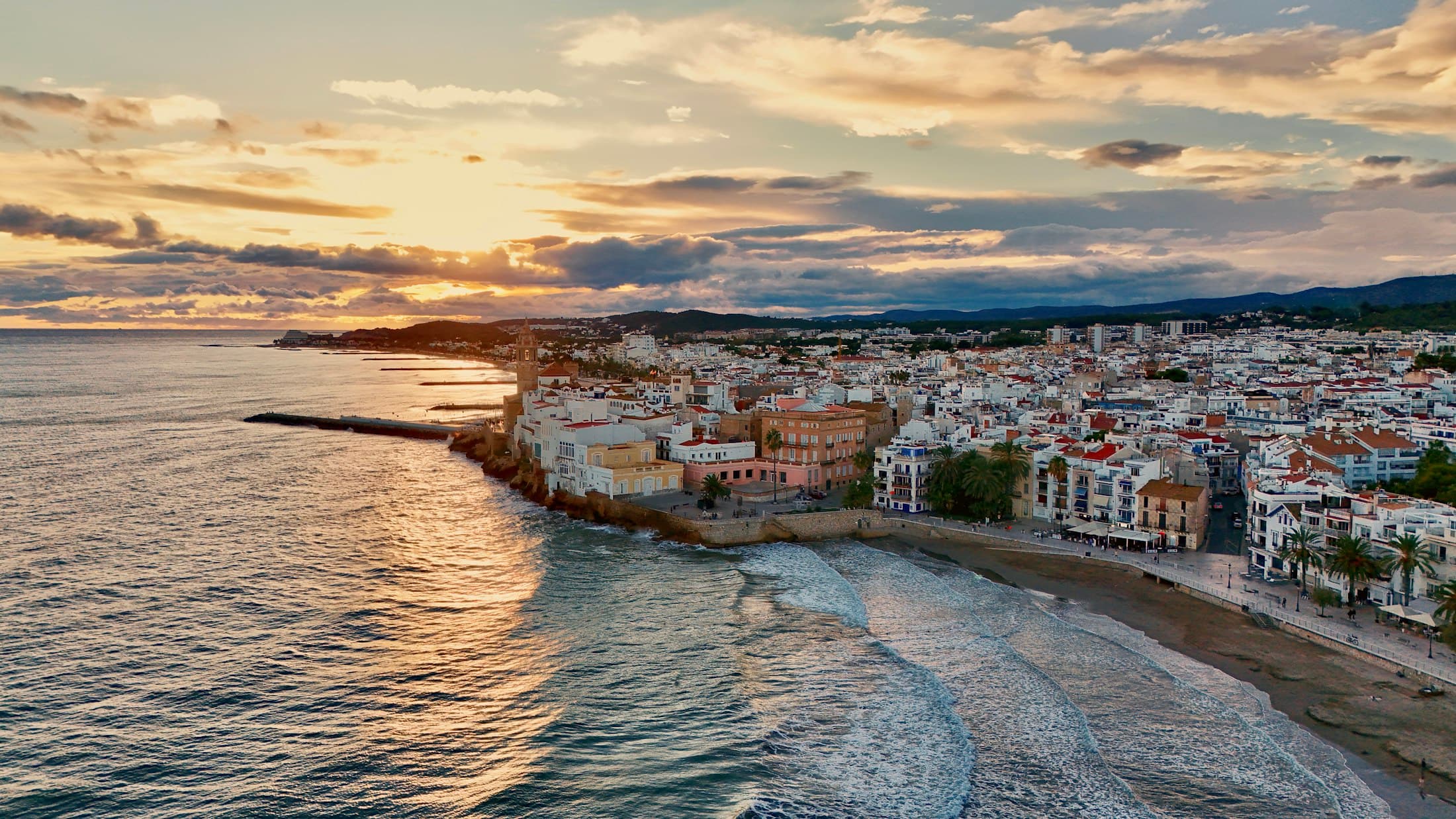 Sitges desde el aire al atardecer: casas blancas y el campanario de la iglesia en la punta, olas que llegan a la playa y las colinas del Garraf detrás