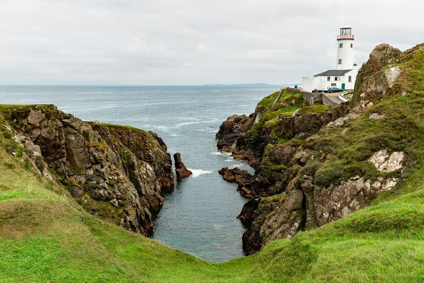 Fanad Head lighthouse and its white keepers’ houses above a narrow rocky inlet, under a grey sky