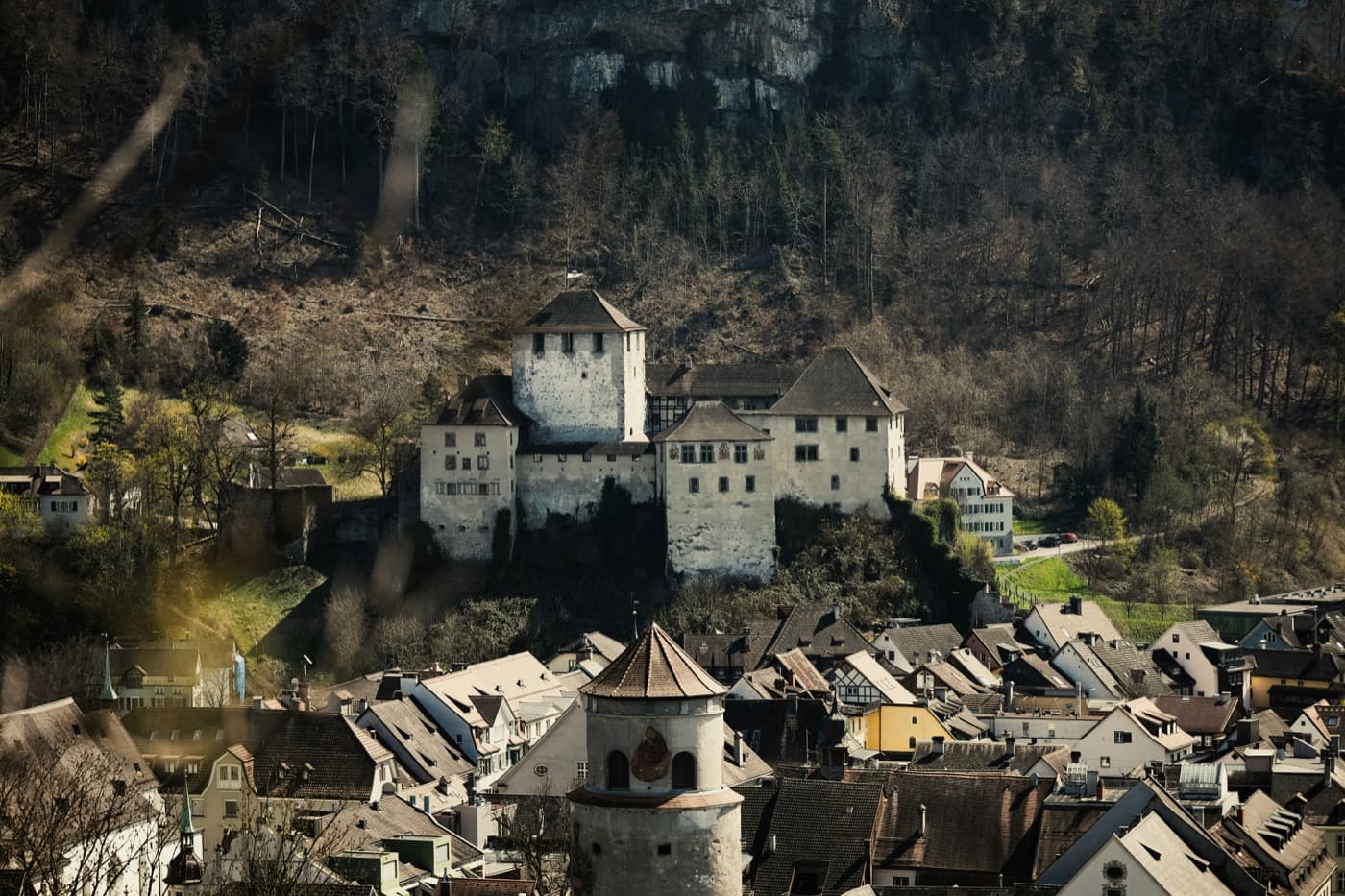 Schattenburg castle, its white keep and walls on a wooded hillside above the old town of Feldkirch, with a round stone tower and steep tiled roofs in front and bare trees on the slope behind