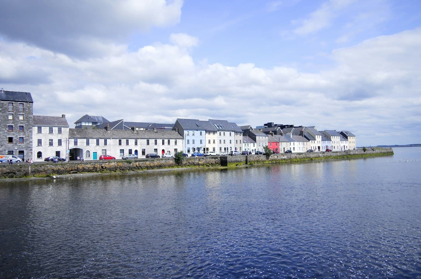 Eine Reihe bunt gestrichener Häuser am steinernen Kai, wo der Corrib auf die Bucht trifft, vom Claddagh aus über das Wasser gesehen