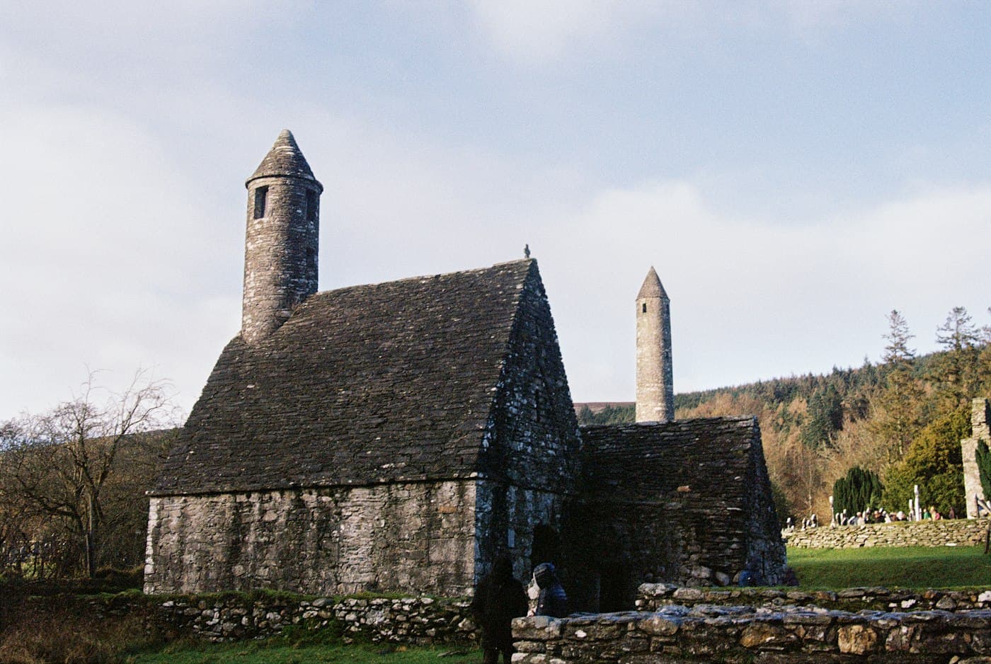 La chiesa di San Kevin a Glendalough, una piccola chiesa di pietra con un ripido tetto di lastre e un campanile rotondo, e la torre rotonda dietro, tra gli alberi