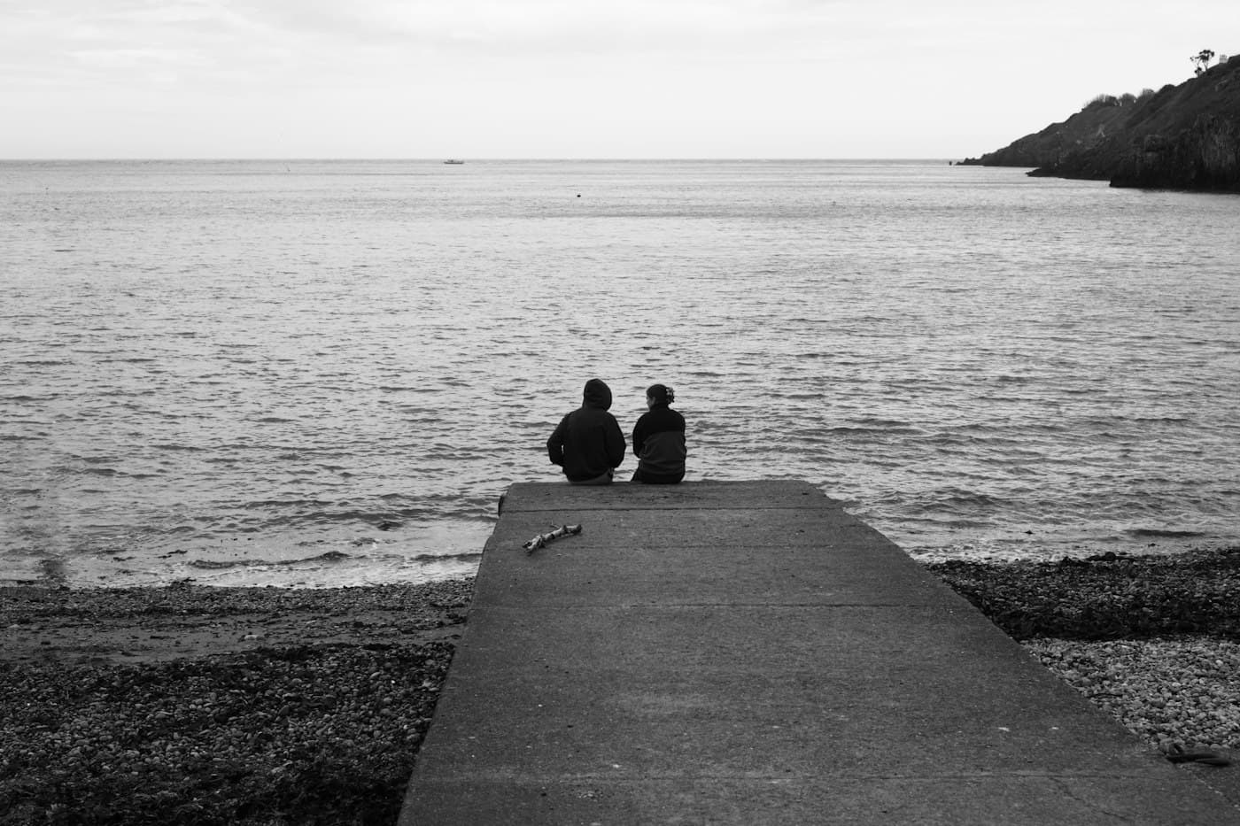 Una coppia seduta in fondo a uno scivolo per le barche a Howth, a guardare un mare grigio e calmo, in bianco e nero