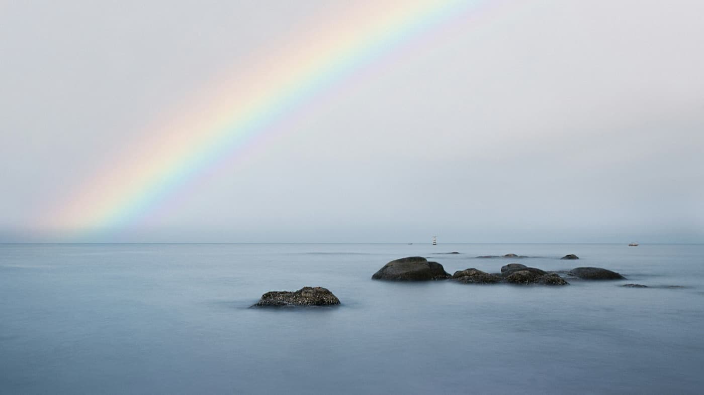 A faint rainbow over a calm, misty sea at Hua Hin, a few dark boulders breaking the smooth water near the shore