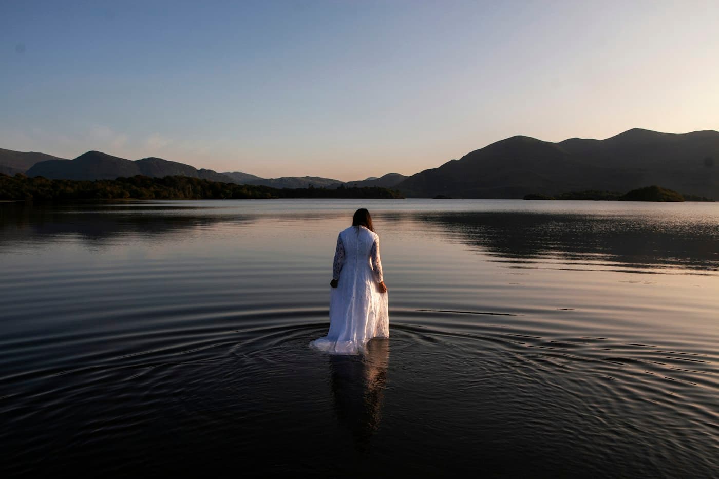 Eine Frau im langen weißen Spitzenbrautkleid watet bei Sonnenuntergang in einen stillen See im Nationalpark Killarney, am anderen Ufer Berge