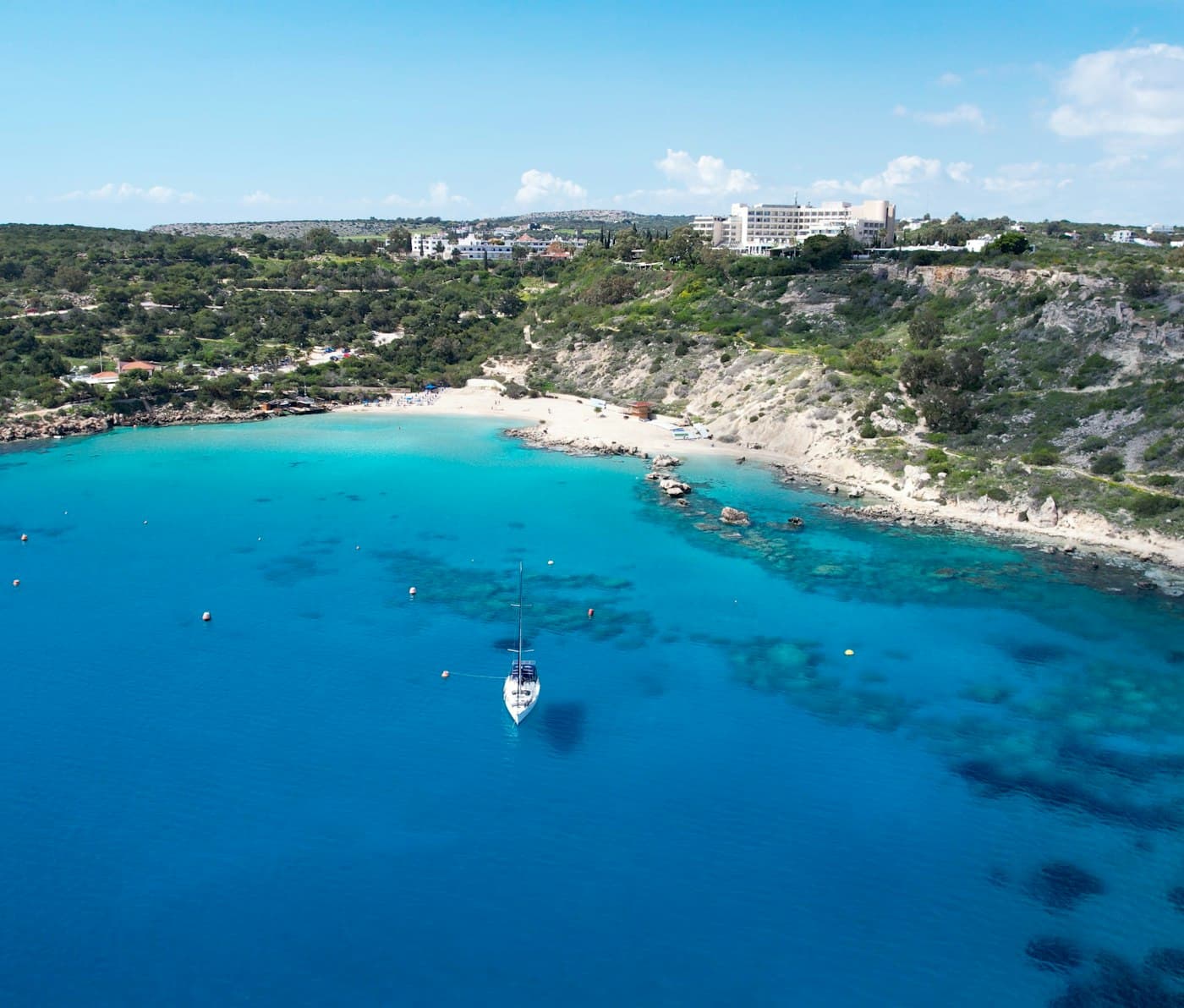 Konnos Bay from the air: a sandy cove of turquoise water with a sailing boat at anchor, below green, scrubby hills