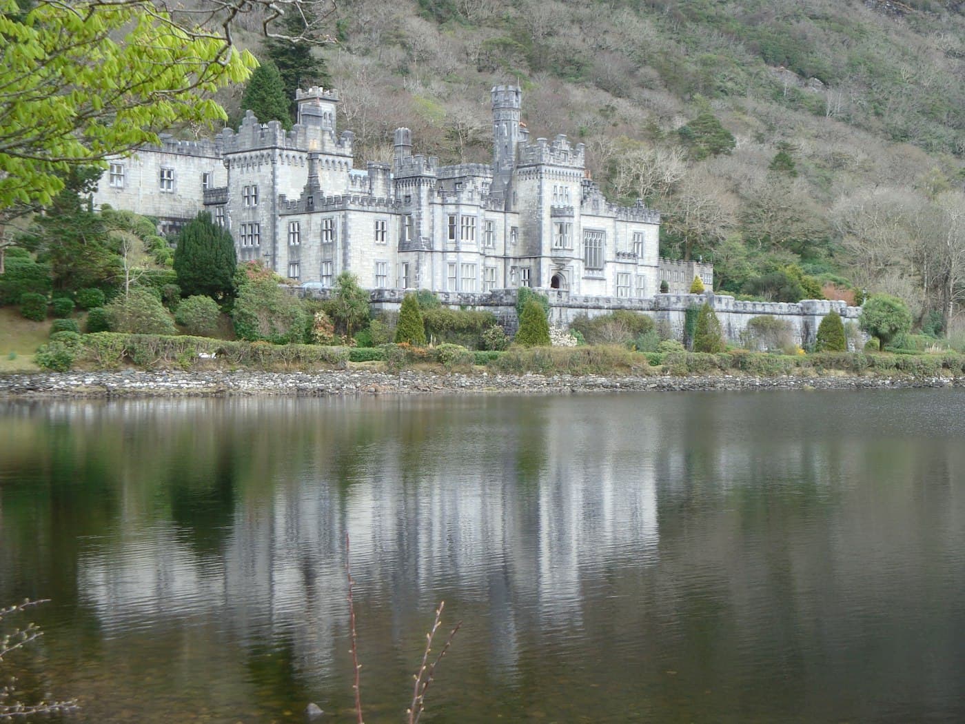 Die graue, zinnenbewehrte Fassade von Kylemore Abbey zwischen Bäumen und Gärten, gespiegelt im See unter einem bewaldeten Berghang