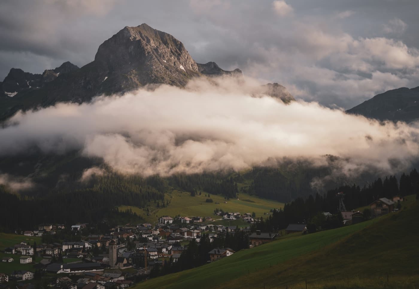 The rocky peak of the Omeshorn rising above a band of cloud, with the village of Lech am Arlberg and its stone church tower in the valley below and a green alpine meadow sloping across the foreground