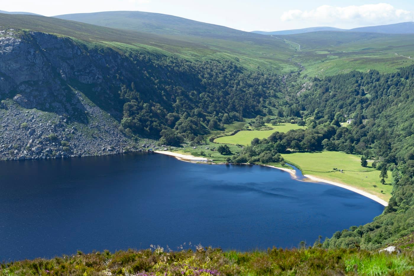 Il Lough Tay, un lago scuro con una riva di sabbia chiara, ai piedi di pendii boscosi e brughiere aperte sui monti Wicklow