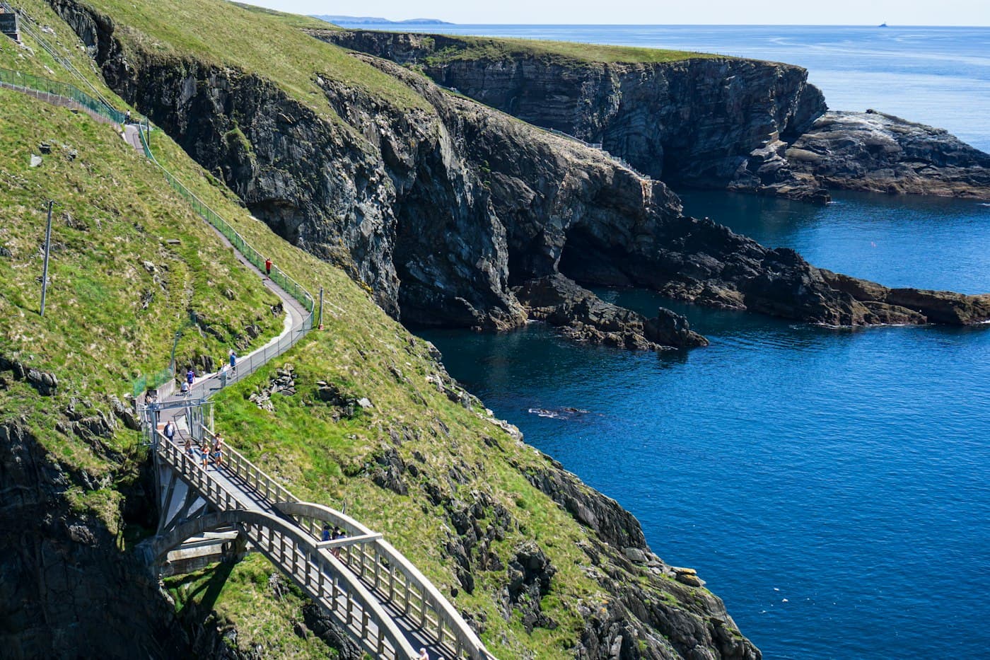 The arched footbridge at Mizen Head above a deep blue inlet, with walkers on the cliff path and dark headlands beyond