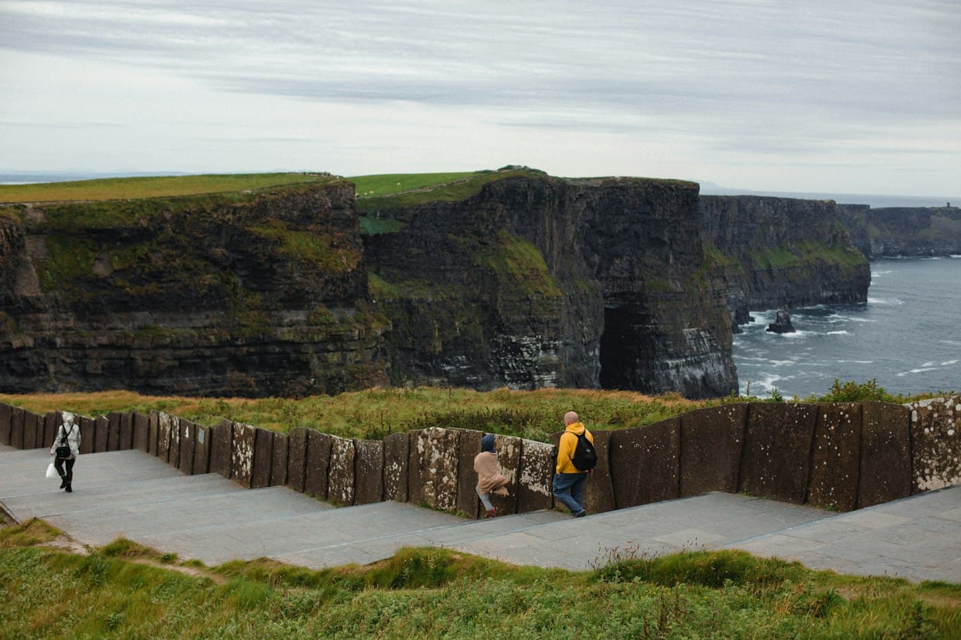 Besucher auf dem gestuften Steinweg hinter der Mauer an den Cliffs of Moher, dahinter die dunklen Klippen und eine Meereshöhle
