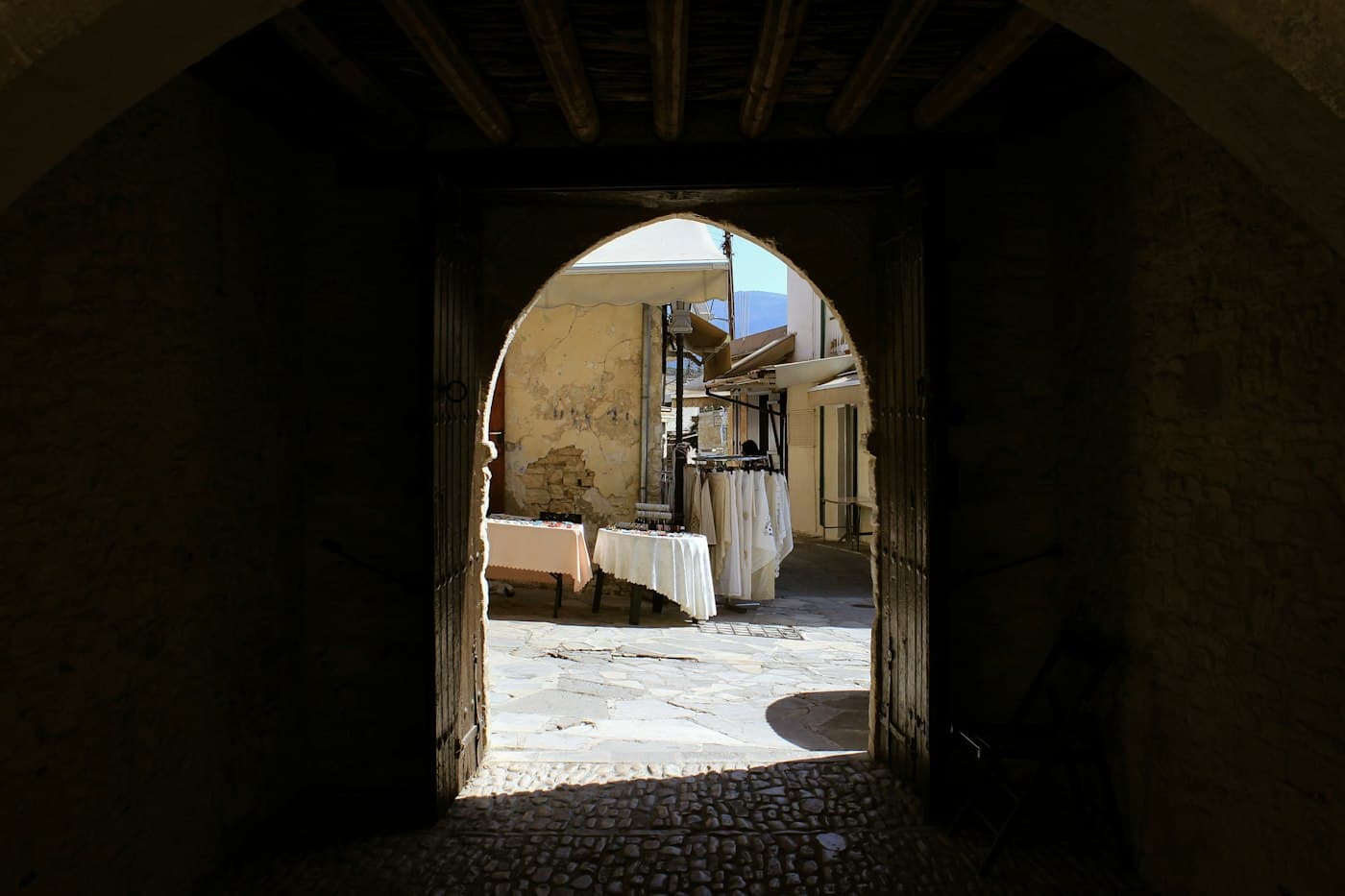 Ein dunkler Steinbogen in Omodos, der sich auf eine sonnige Gasse mit Tischen und Ständern voller weißer Spitze und Leinen öffnet