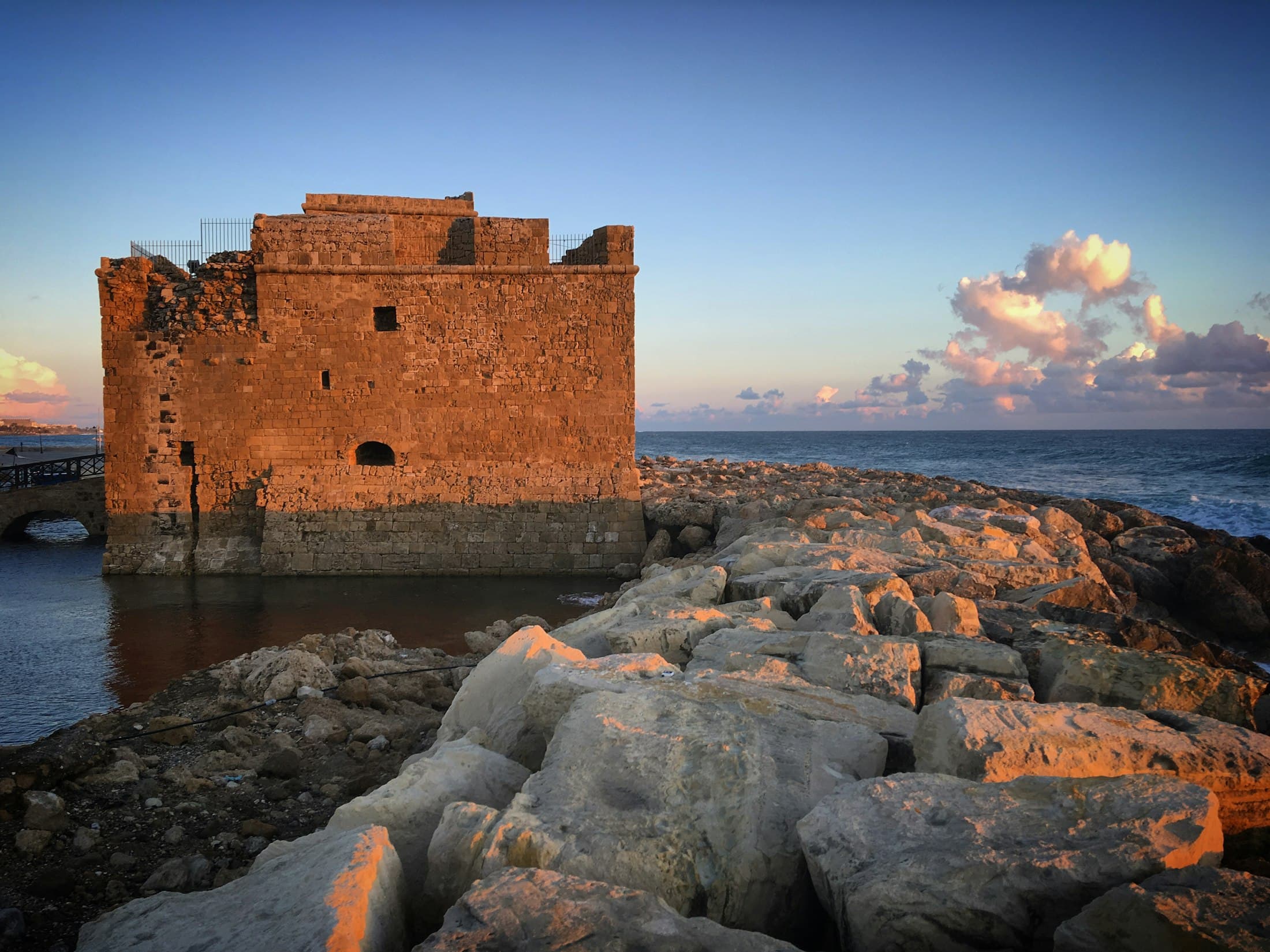 Das steinerne Fort am Ende des Hafens von Paphos, orange in der Abendsonne, davor die hellen Felsen der Mole, dahinter das Meer