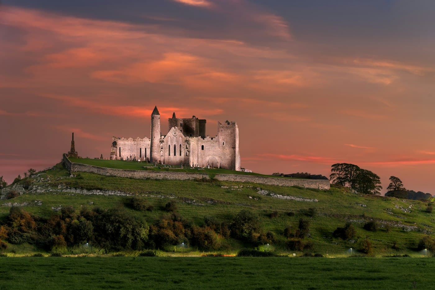 La Rocca di Cashel illuminata sulla sua collina verde sotto un cielo rosso di sera, con la torre rotonda e la cattedrale in cima