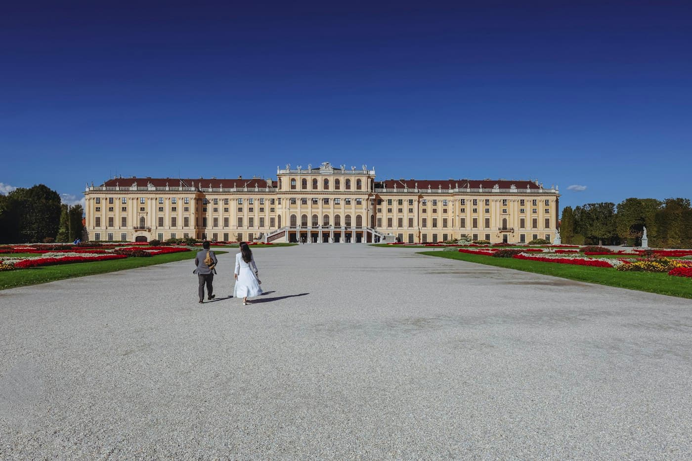 Ein Paar, sie im weißen Kleid, geht über den Kies des Schönbrunner Schlossparks auf das Schloss zu